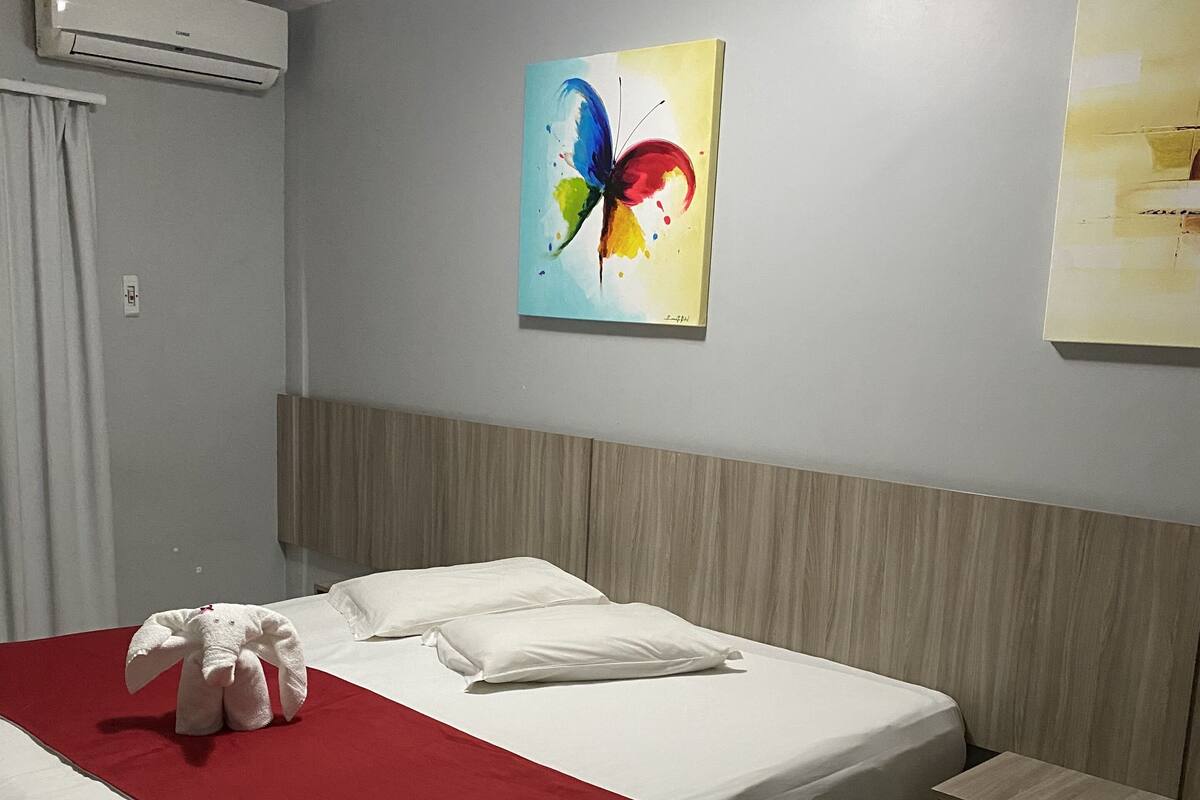 A hotel room with a bed, a red blanket, a white pillow, a wooden nightstand, and an air conditioning unit.
