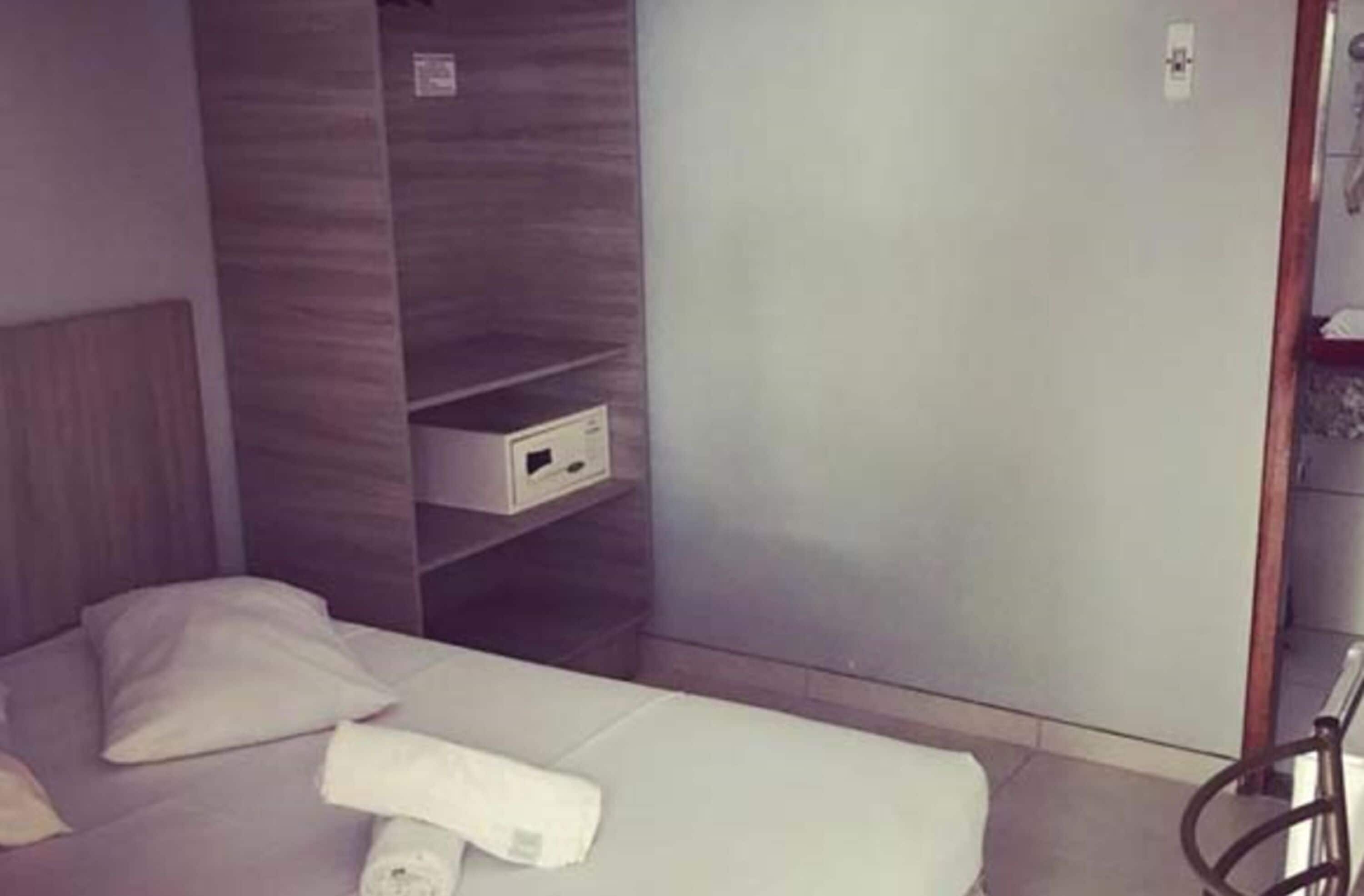 Une chambre d’hôtel avec un lit, une petite table de chevet blanche, une étagère fixée au mur et une tête de lit en bois.