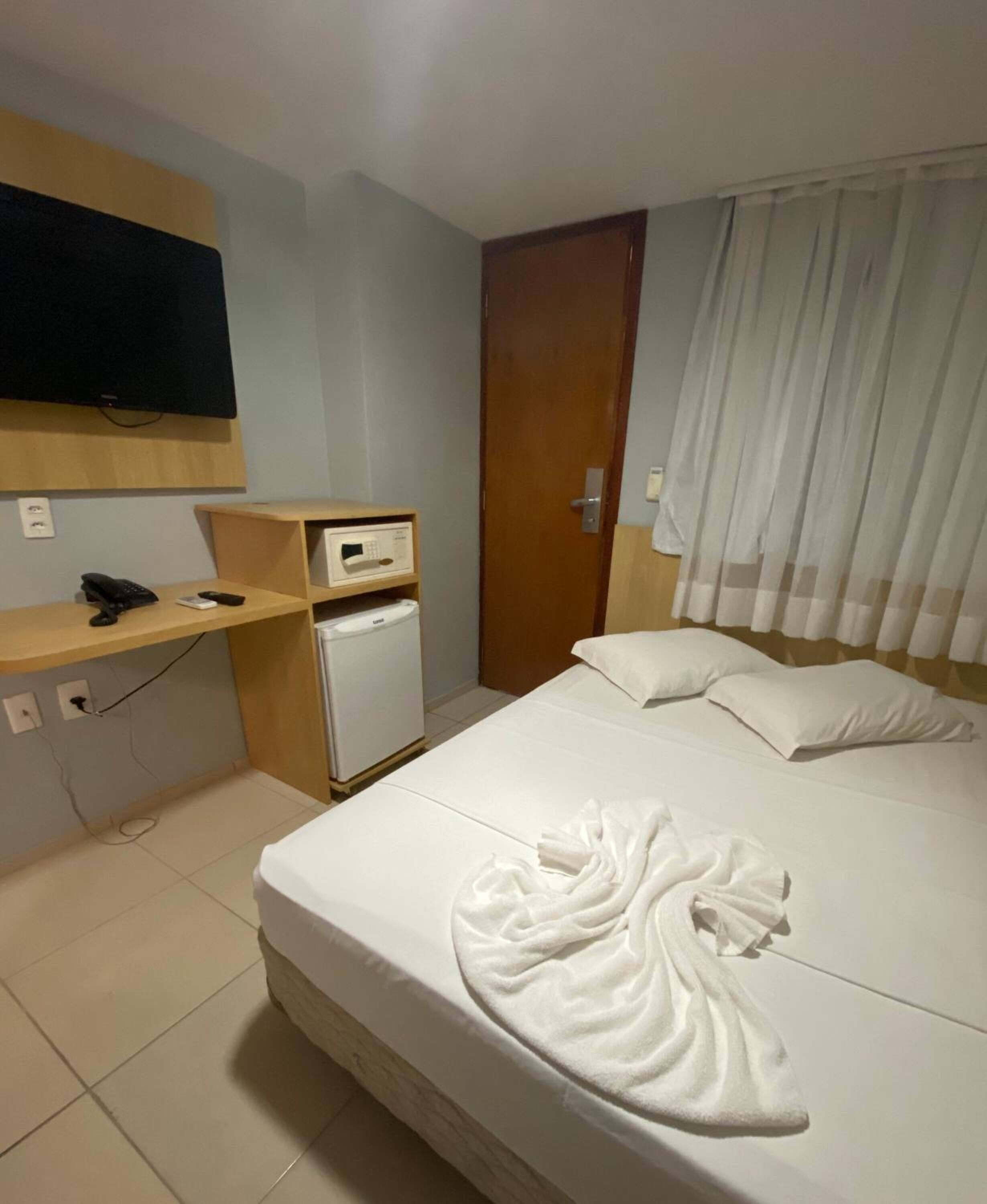 A hotel room with a bed, a TV mounted on the wall, a small refrigerator, and a wooden shelf with a phone and remote control.