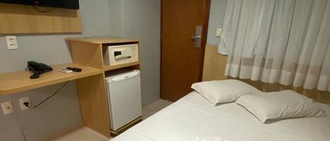 Habitación de hotel con cama, televisión montada en la pared, refrigerador pequeño y estante de madera con teléfono y control remoto.