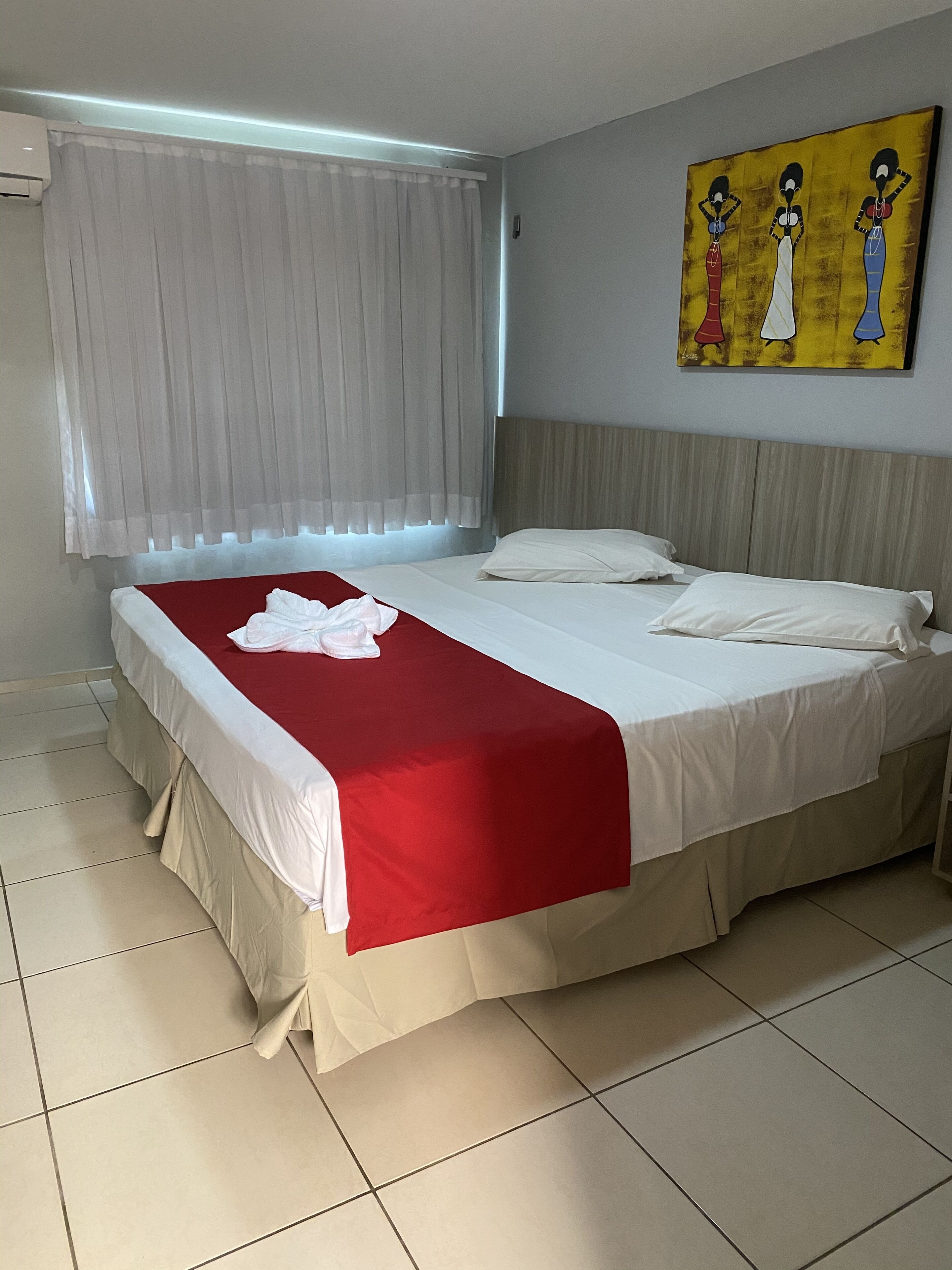 Una habitación de hotel con cama, cortinas blancas, un cuadro en la pared y suelo de baldosas.