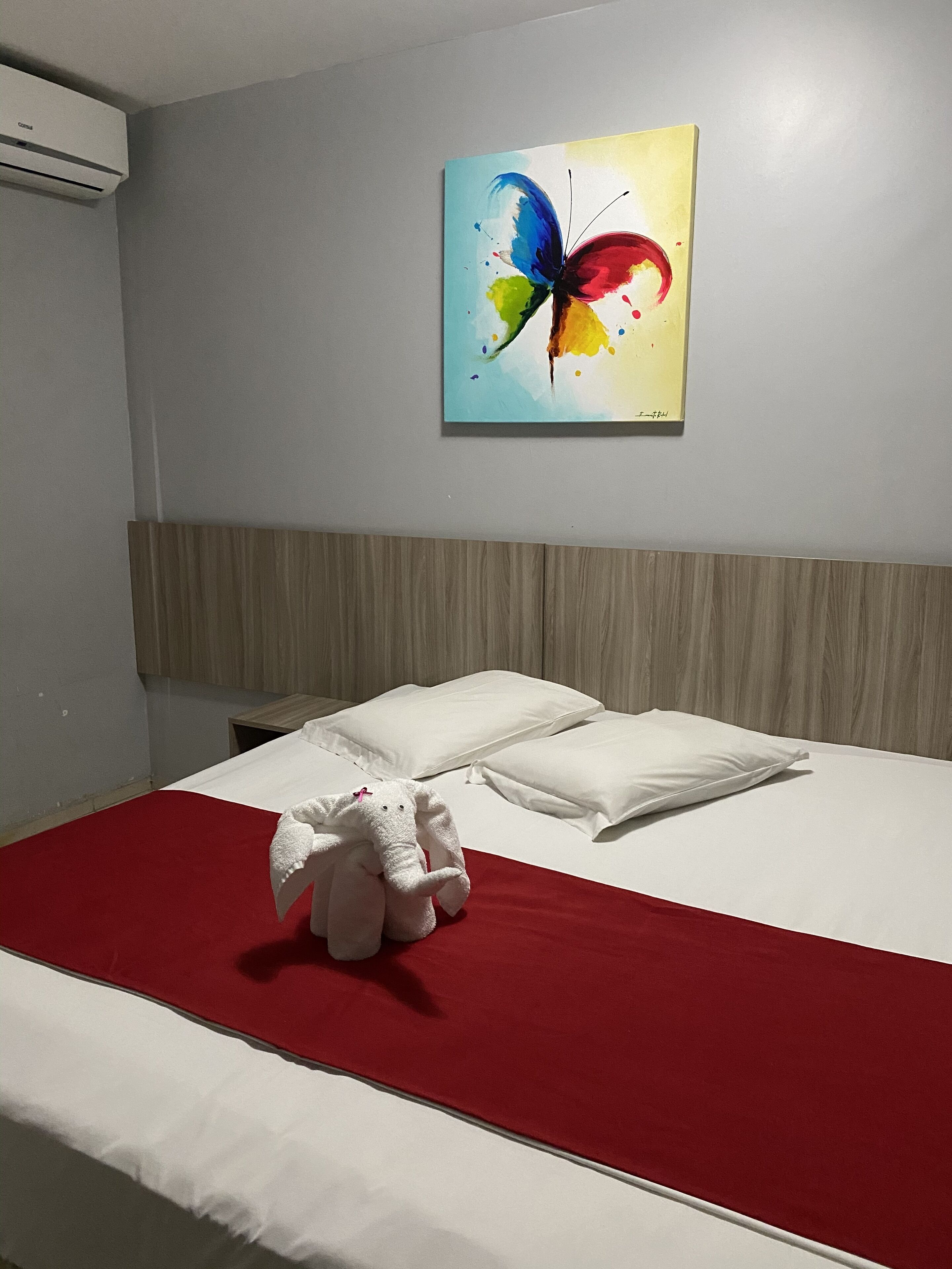 A hotel room with a bed, white pillows, a red bedspread, and a colorful butterfly painting on the wall.