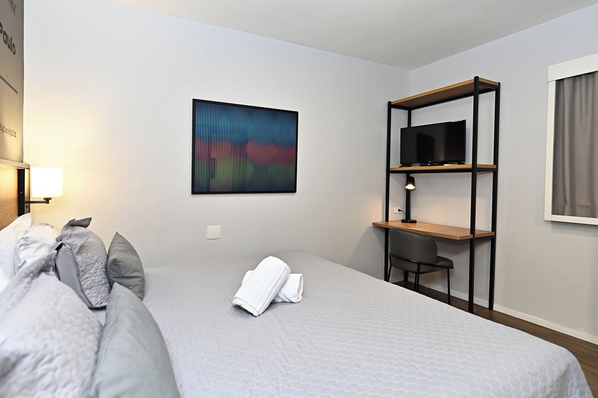 Una camera d'albergo con un letto, una televisione su un ripiano, una lampada e un quadro appeso al muro.