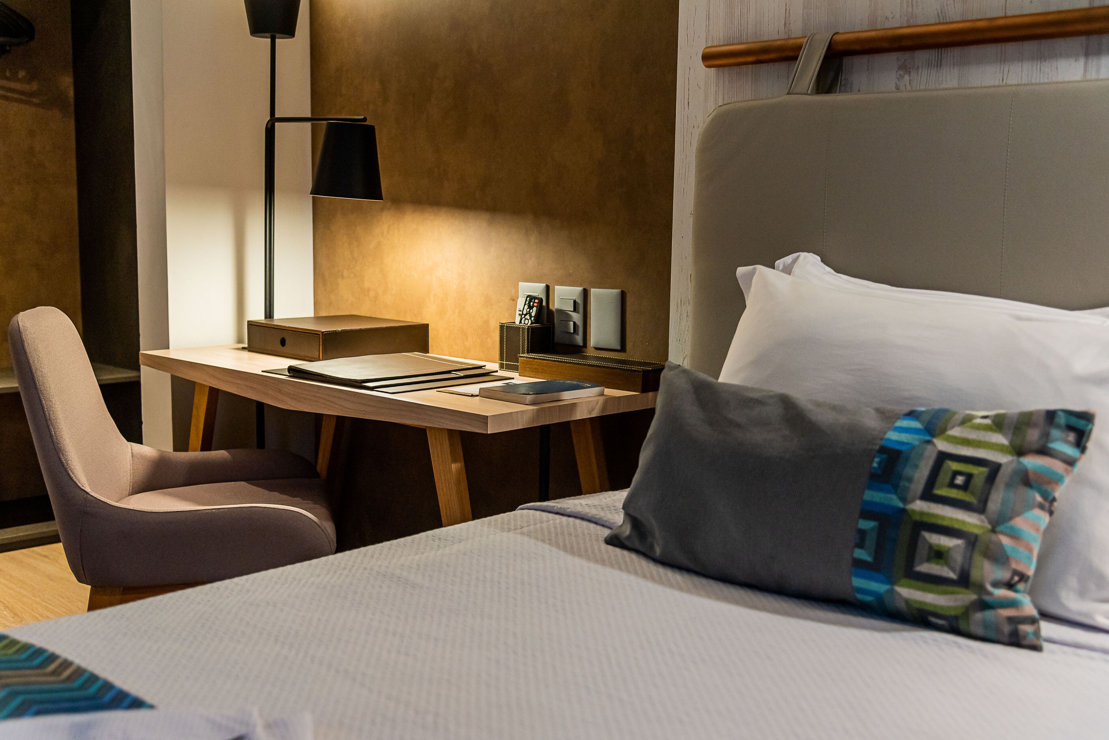 Quarto de hotel com cama, escrivaninha, cadeira, abajur e mesa de cabeceira.