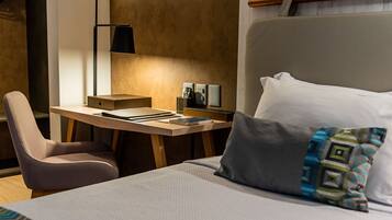 Quarto de hotel com cama, escrivaninha, cadeira, abajur e mesa de cabeceira.