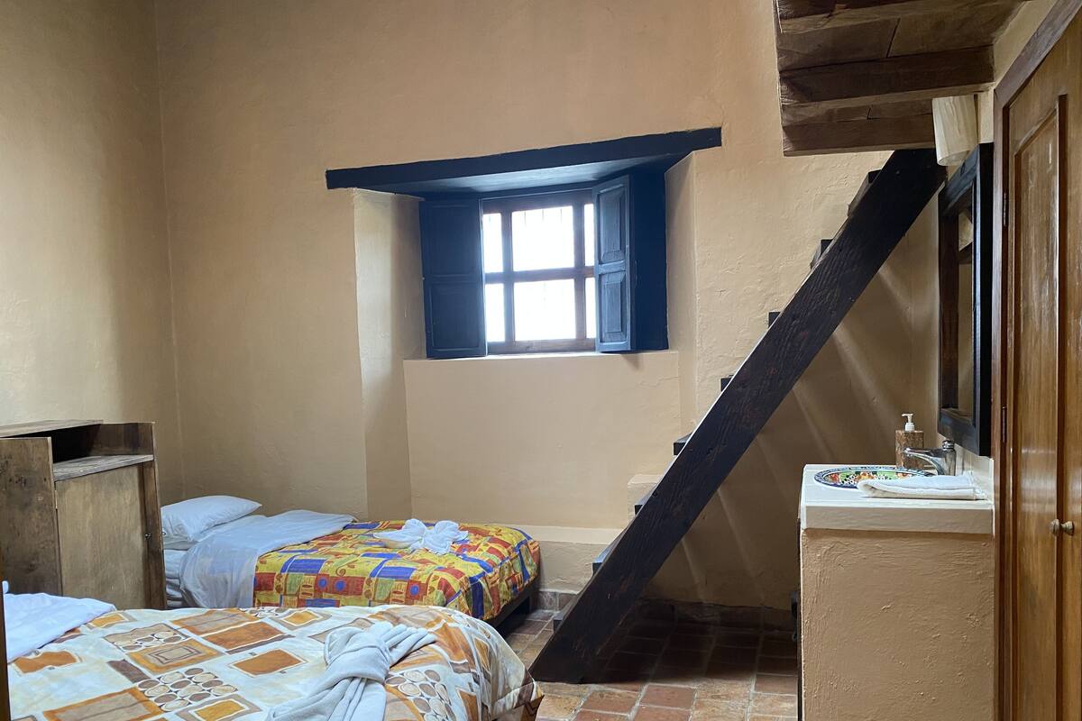 Une petite pièce agréable, dotée de deux lits, d’une fenêtre, d’un escalier en bois et d’un lavabo avec un miroir.