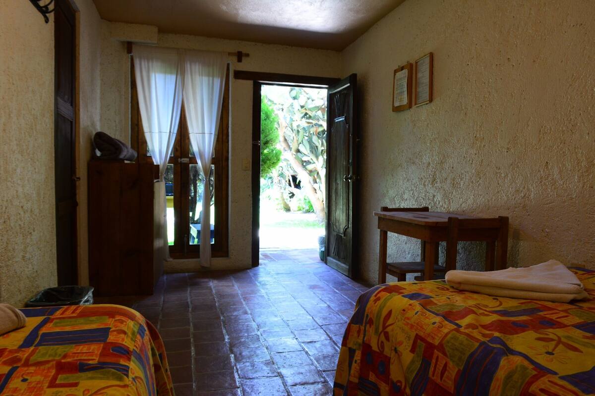 Une chambre avec deux lits, une table en bois et une vue sur un jardin, accessible par une porte ouverte.