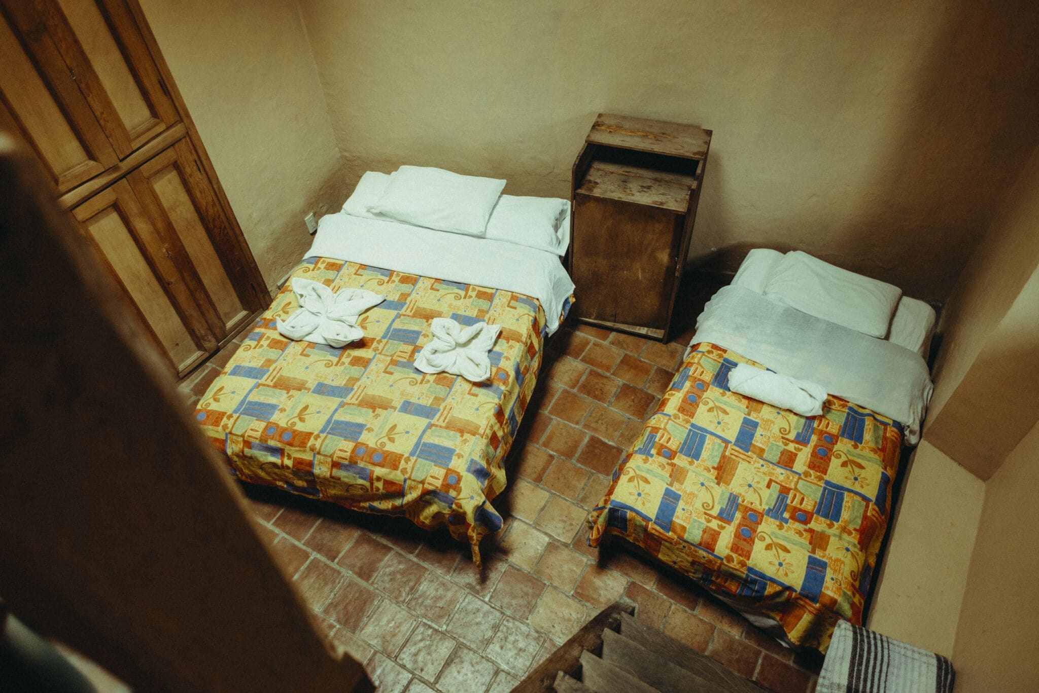 Dos camas con colchas estampadas y ropa de cama blanca, una mesita de noche de madera y un suelo de baldosas.
