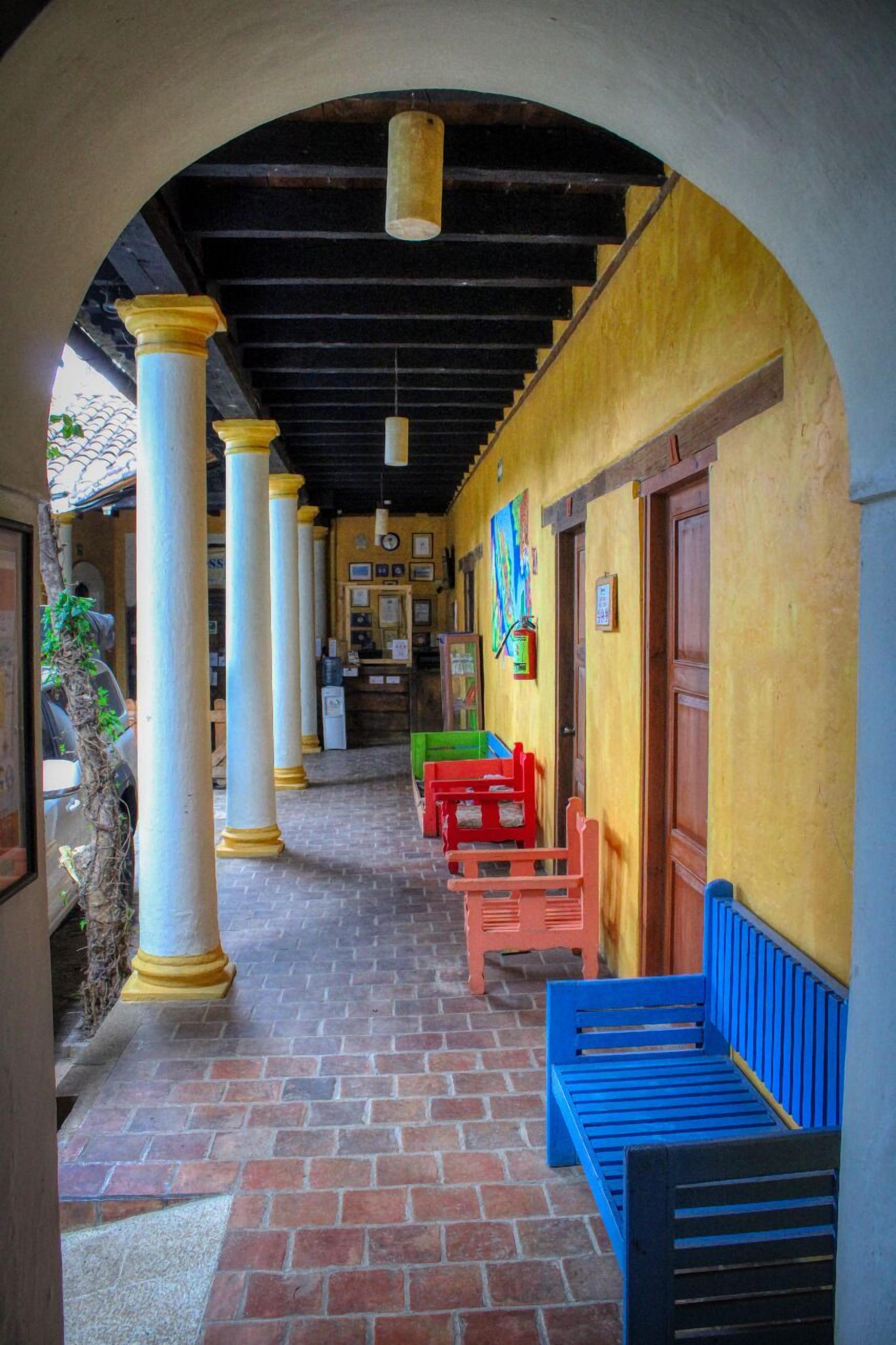 A covered corridor with columns, benches, and a brick floor.