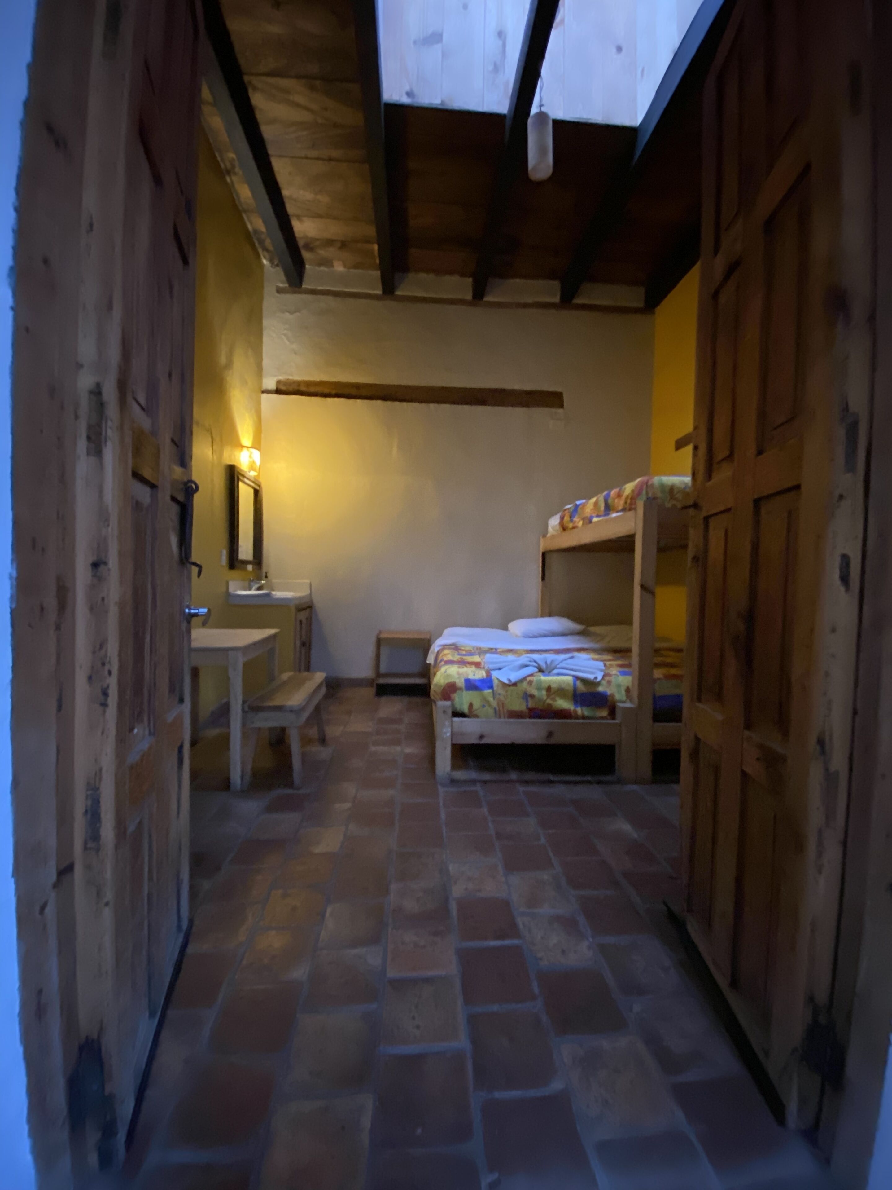 Un pasillo estrecho que conduce a una habitación con una cama, una mesita pequeña y un banco. La habitación tiene paredes de madera y suelo de baldosas.