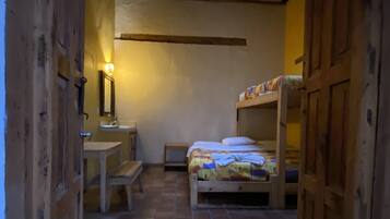 Un pasillo estrecho que conduce a una habitación con una cama, una mesita pequeña y un banco. La habitación tiene paredes de madera y suelo de baldosas.