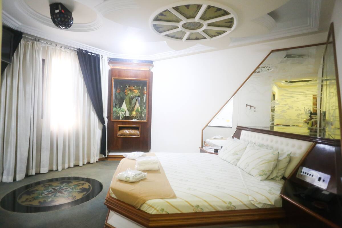 Una habitació d'hotel amb un llit gran, un amb motllures de fusta, una làmpada de sostre rodona, un mirall i una finestra amb cortines.