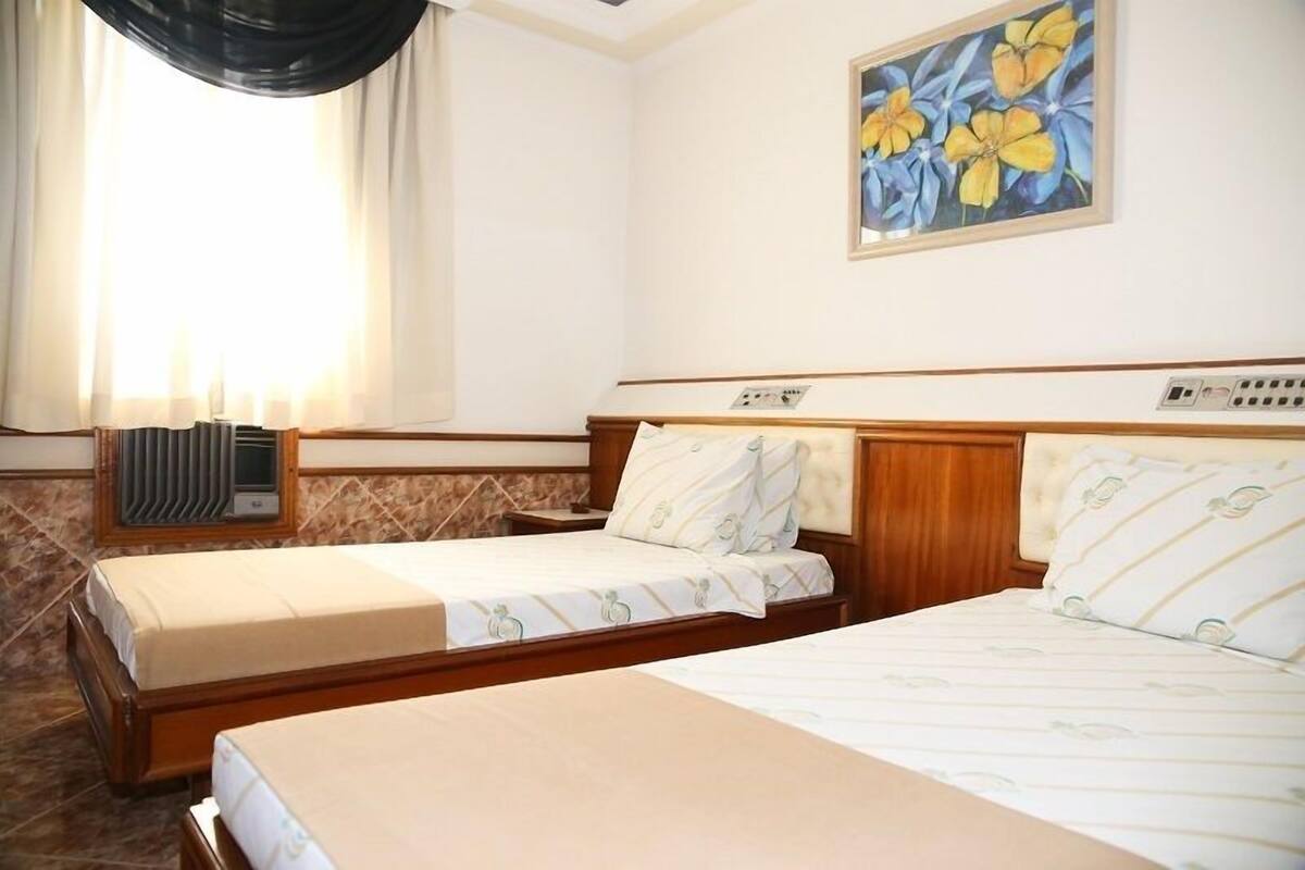 Habitació d'hotel amb dos llits, una finestra amb cortines, un radiador i un quadre de flors a la paret.