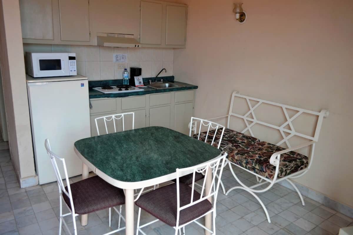 A compact kitchen with a microwave, sink, and a small refrigerator. There is a dining table with four chairs and a bench with a floral cushion.