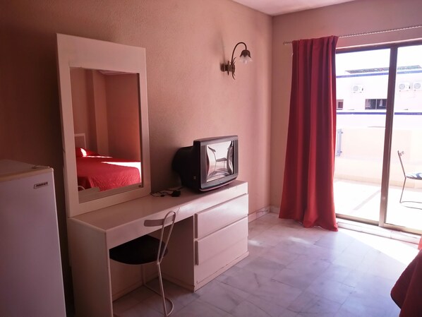 A hotel room with a bed, a desk with a television, a mirror, a small refrigerator, and a sliding glass door with a red curtain. - Los Jitos Hotel & Suites (San Carlos)