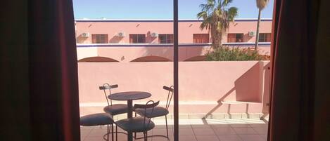 A room with a sliding glass door leading to an outdoor patio. The patio has a tiled floor and a round table with chairs. There is a palm tree and a building with a pink facade in the background.