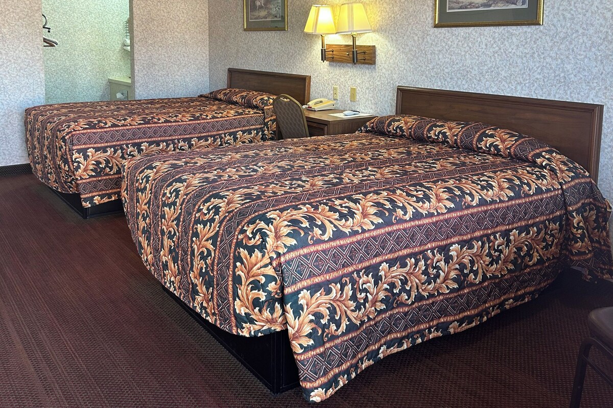 A hotel room with two beds, a desk, a chair, a lamp, and two framed pictures on the wall.