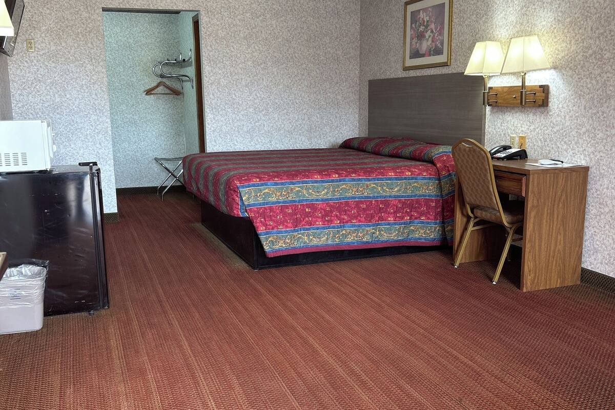 A hotel room with a bed, desk, chair, and a small table with a lamp.