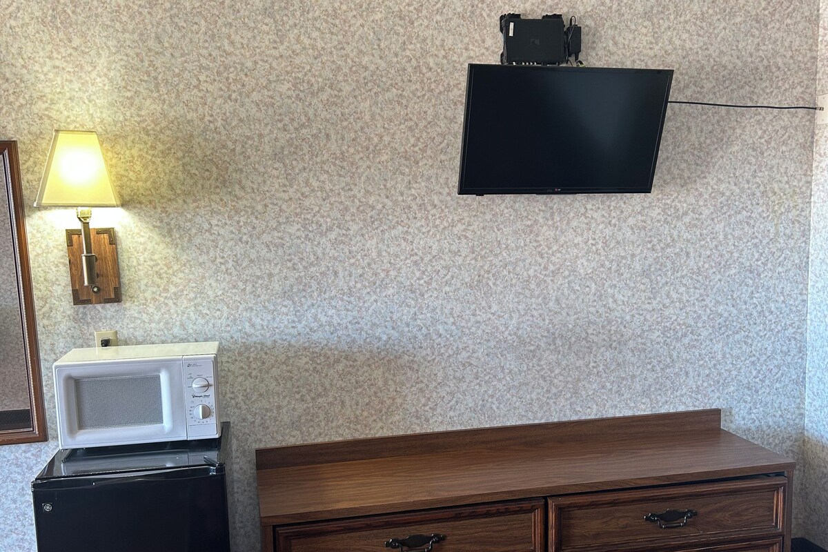A hotel room with a bed, a wooden dresser, a microwave, and a flat-screen TV mounted on the wall.
