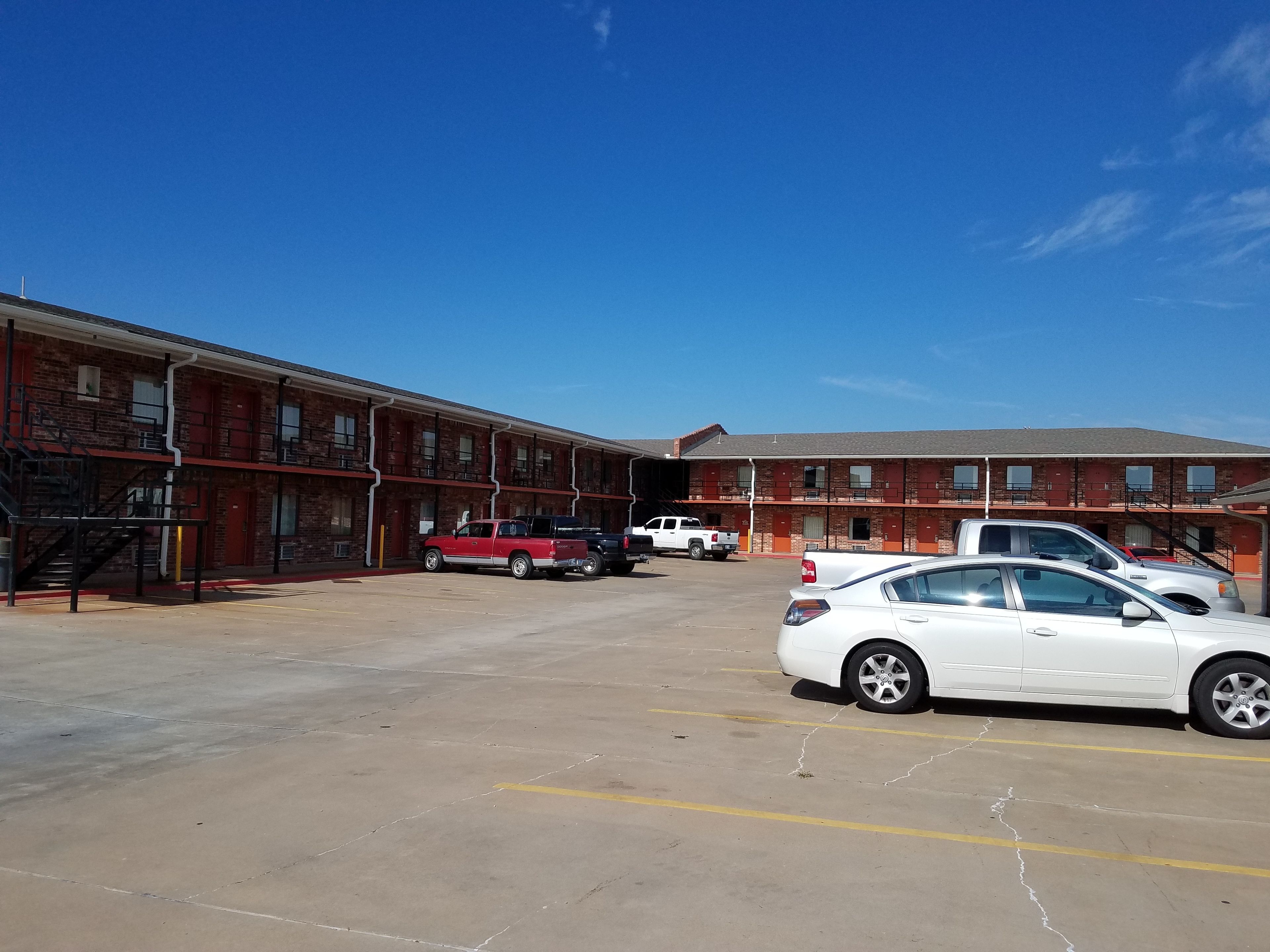 Un motel à plusieurs étages, avec une façade en briques, un parking où sont garées plusieurs voitures et un ciel d’un bleu limpide.