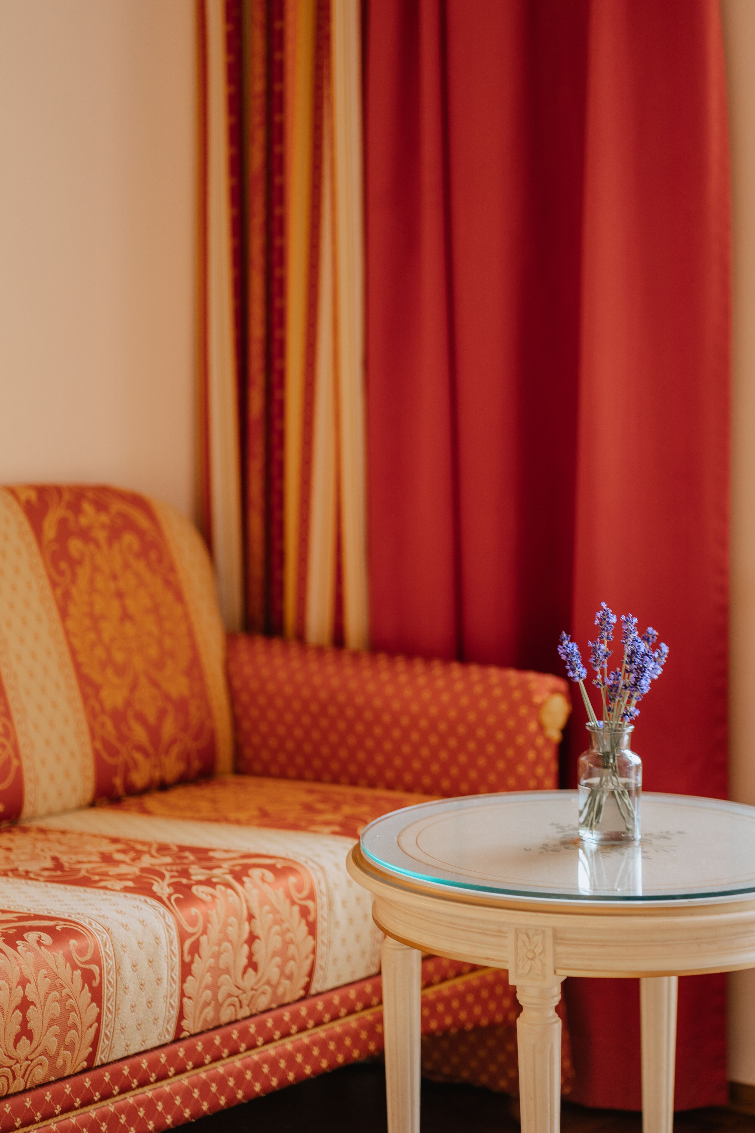 A room with a patterned sofa, a small round table with a vase of flowers, and red curtains.
