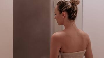 A person standing in a bathroom with a towel wrapped around their body, a glass shower door, and a plain wall.
