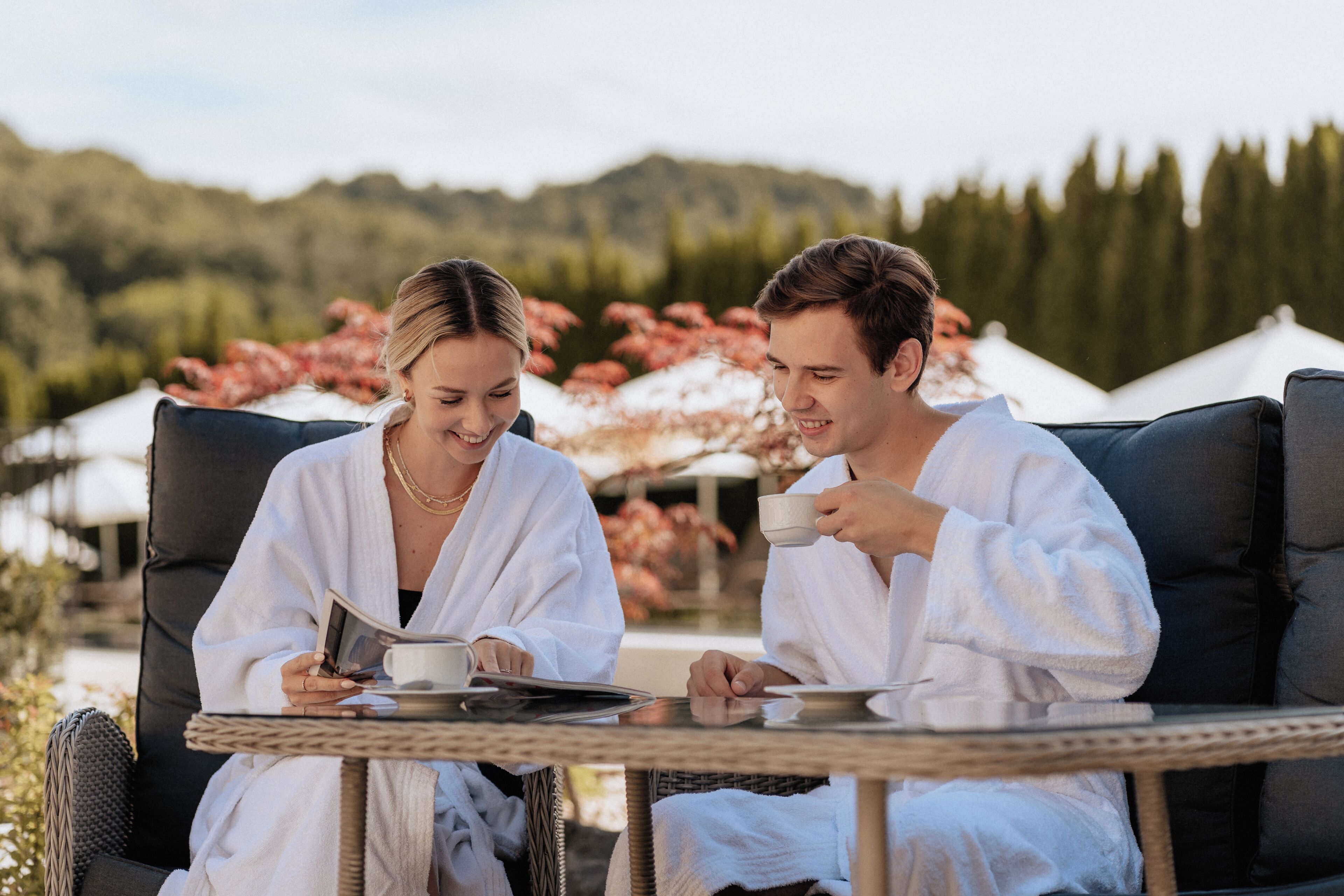 Zwei Personen in weißen Roben sitzen an einem Tisch im Freien mit Blick auf Bäume und Hügel im Hintergrund.