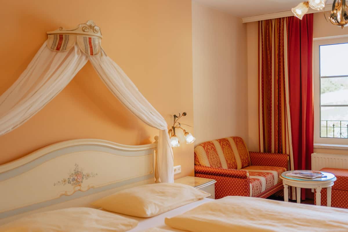 Un dormitorio con cama con dosel, un sofá, una mesita y una ventana con cortinas.