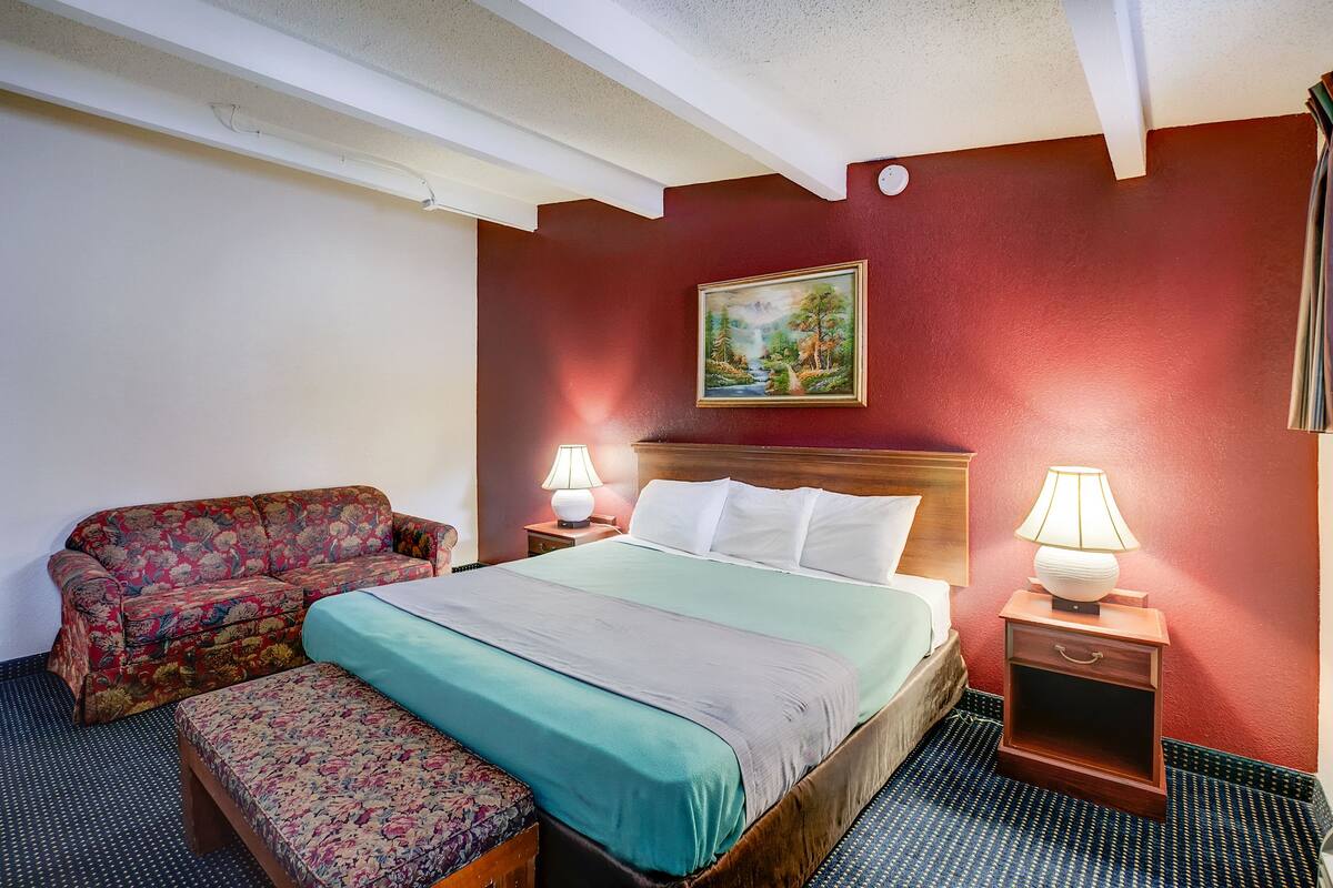 A hotel room with a bed, a sofa, a nightstand with a lamp, and a painting on the wall.