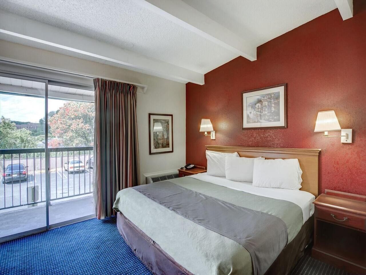 A hotel room with a large bed, two bedside lamps, a framed picture on the wall, and a view of a parking area through a sliding door.