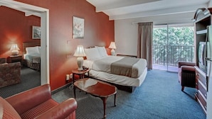 Ein Hotelzimmer mit einem Bett, zwei Nachttischen mit Lampen, einem Sofa, einem kleinen Tisch, einem Fernseher und einem Balkon mit Blick auf Bäume.