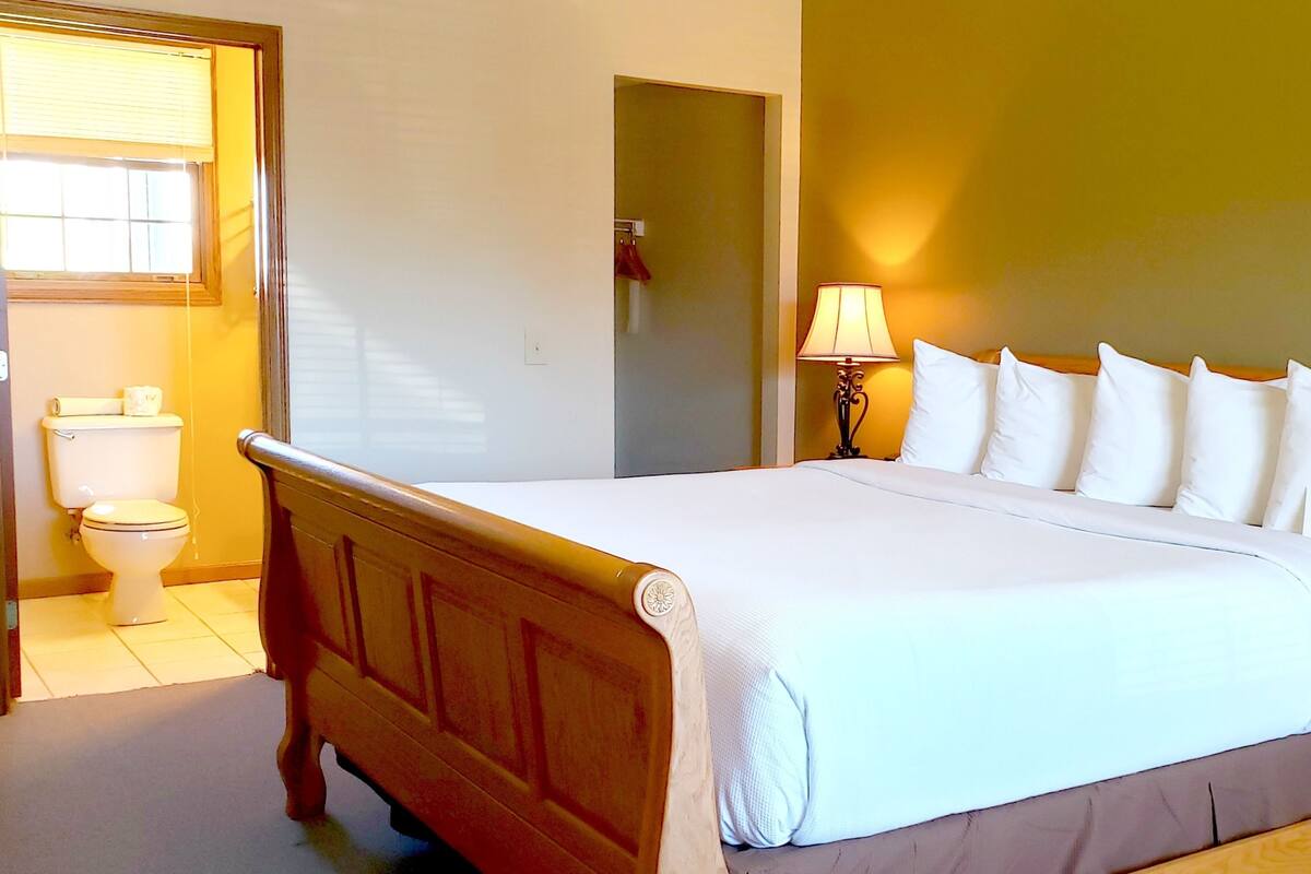 A neatly made bed with white bedding and a wooden headboard, a bedside lamp, and a toilet in a room with yellow walls.