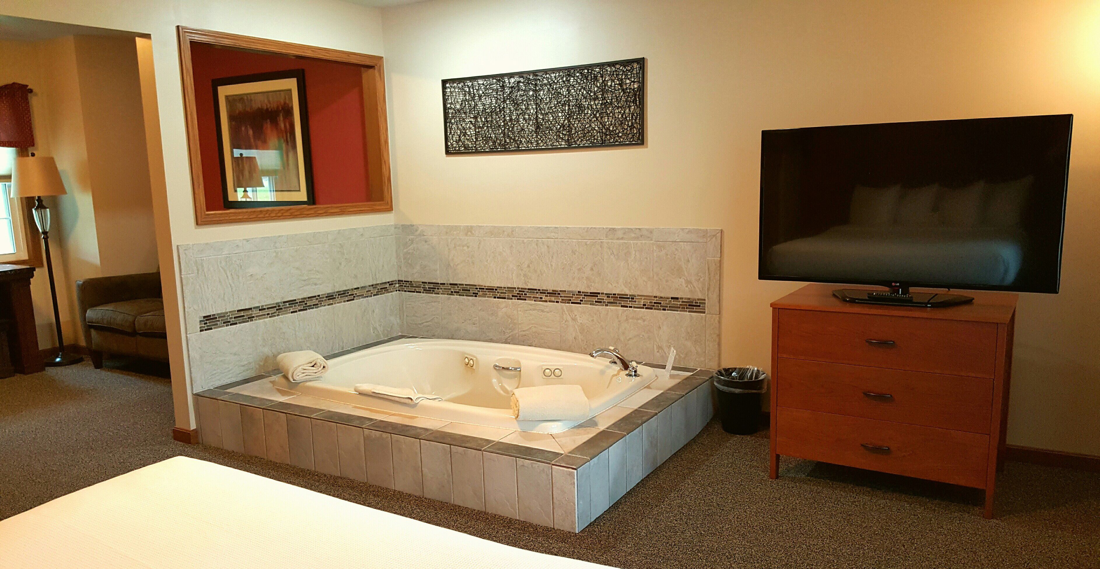 A hotel room with a large bathtub, a television on a wooden dresser, and a mirror above the bathtub.