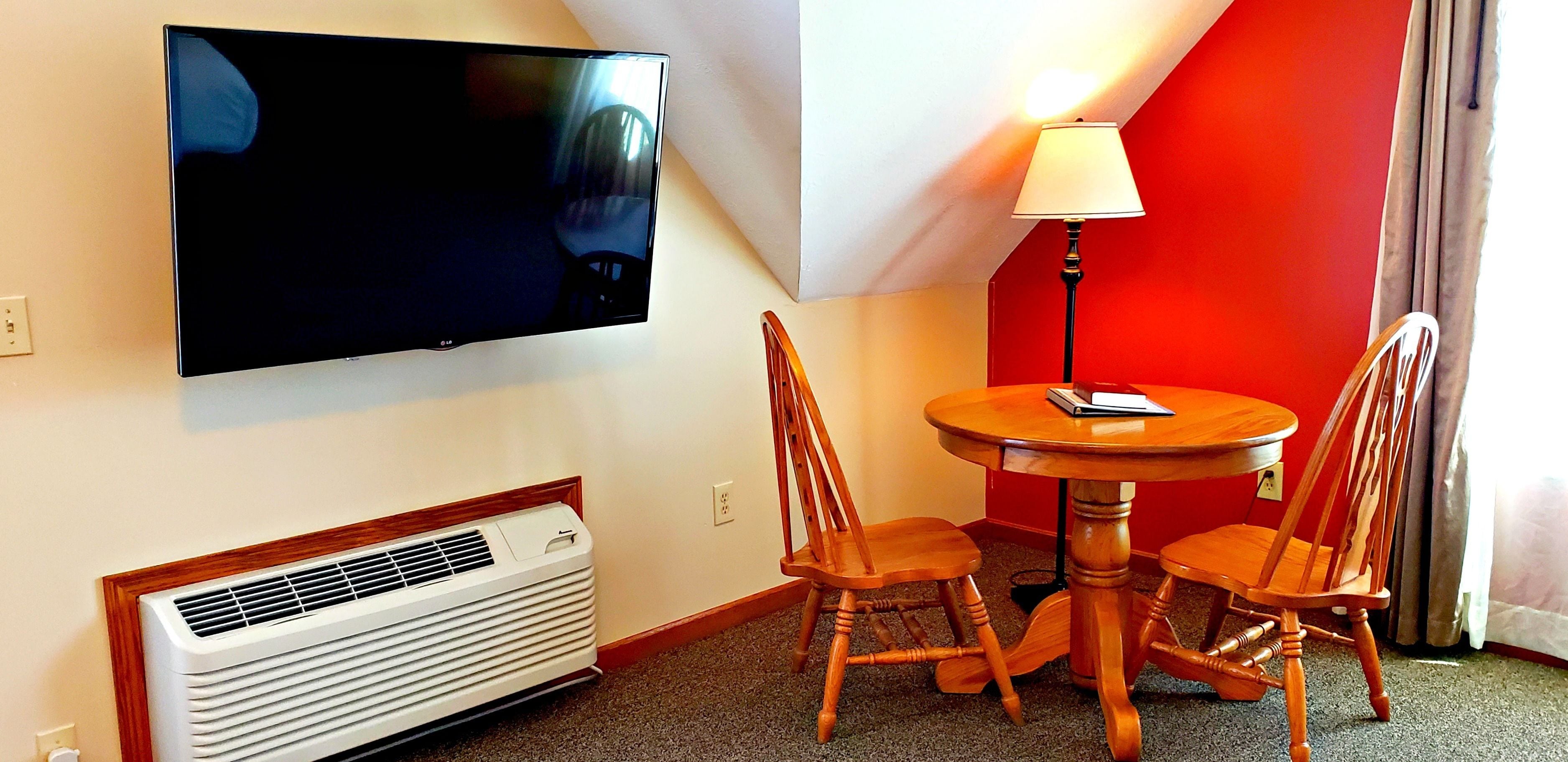 A hotel room with a flat-screen TV mounted on the wall, a round wooden table with two chairs, a standing lamp, and a wall-mounted heater.