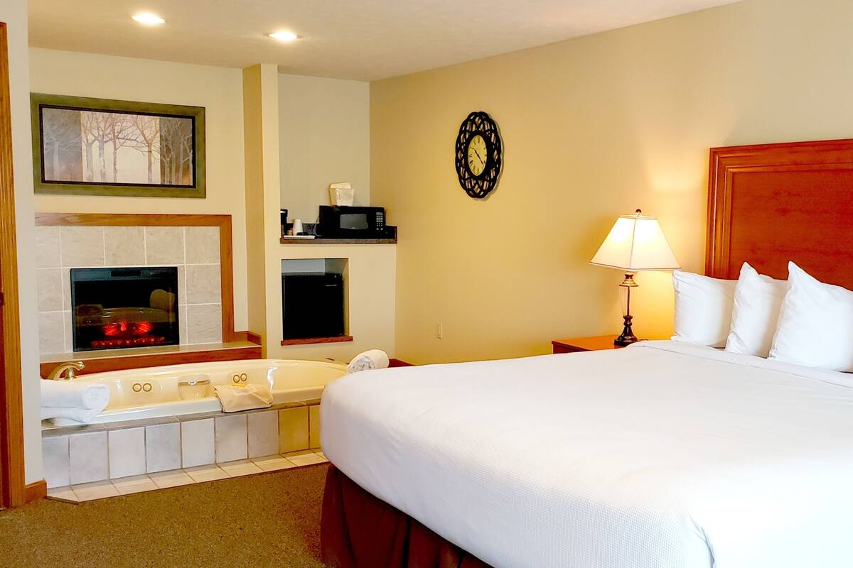 A hotel room with a large bed, a bathroom with a bathtub, a microwave, a clock, and a fireplace.