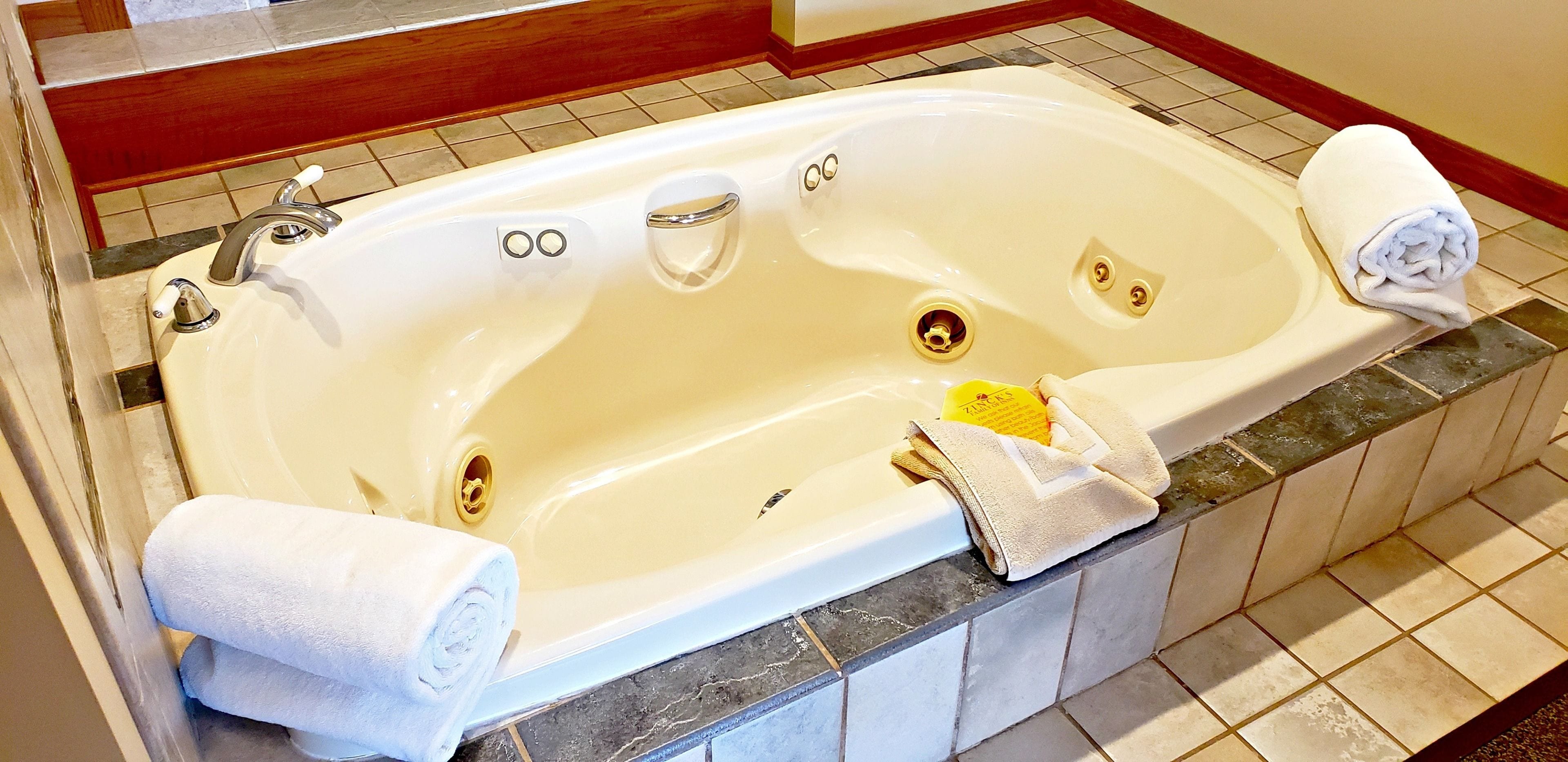 A large bathtub with two faucets, a drain, and a yellow bottle. There are two rolled towels on the edge.