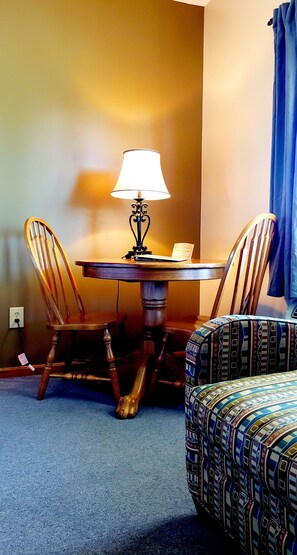 A room with a round wooden table, two wooden chairs, a patterned sofa, and a lamp.