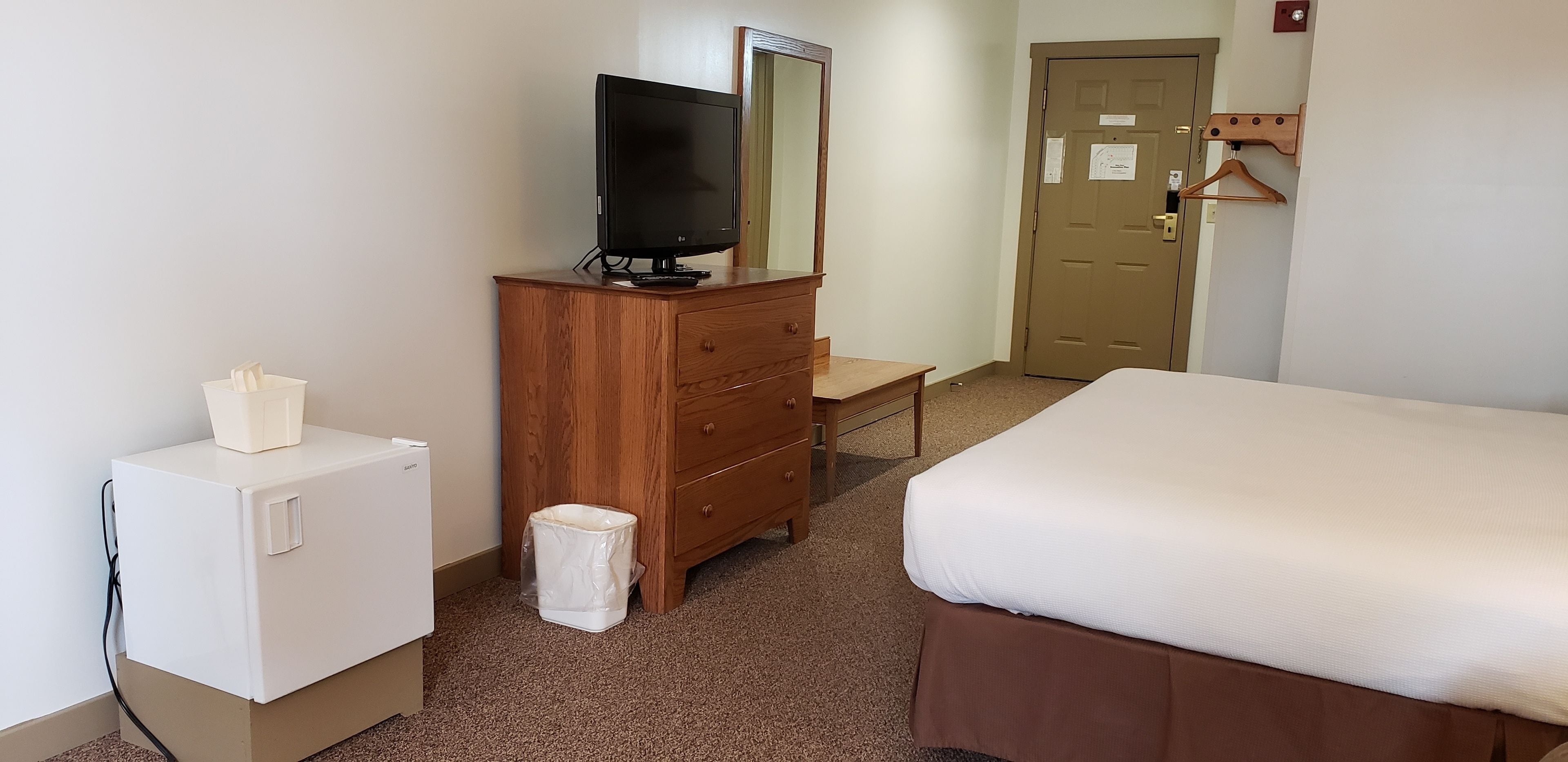 Una habitación de hotel con un aparador de madera, un televisor, un refrigerador blanco pequeño, una cama y una puerta con un perchero de madera.