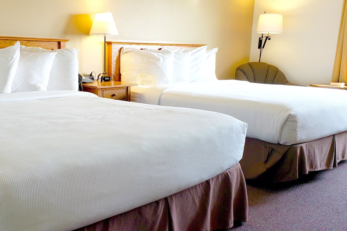 A hotel room with two beds, each with white linens and a wooden headboard. There are bedside tables with lamps and a small clock.