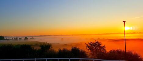 A sunrise over a misty landscape with a road and a white fence.