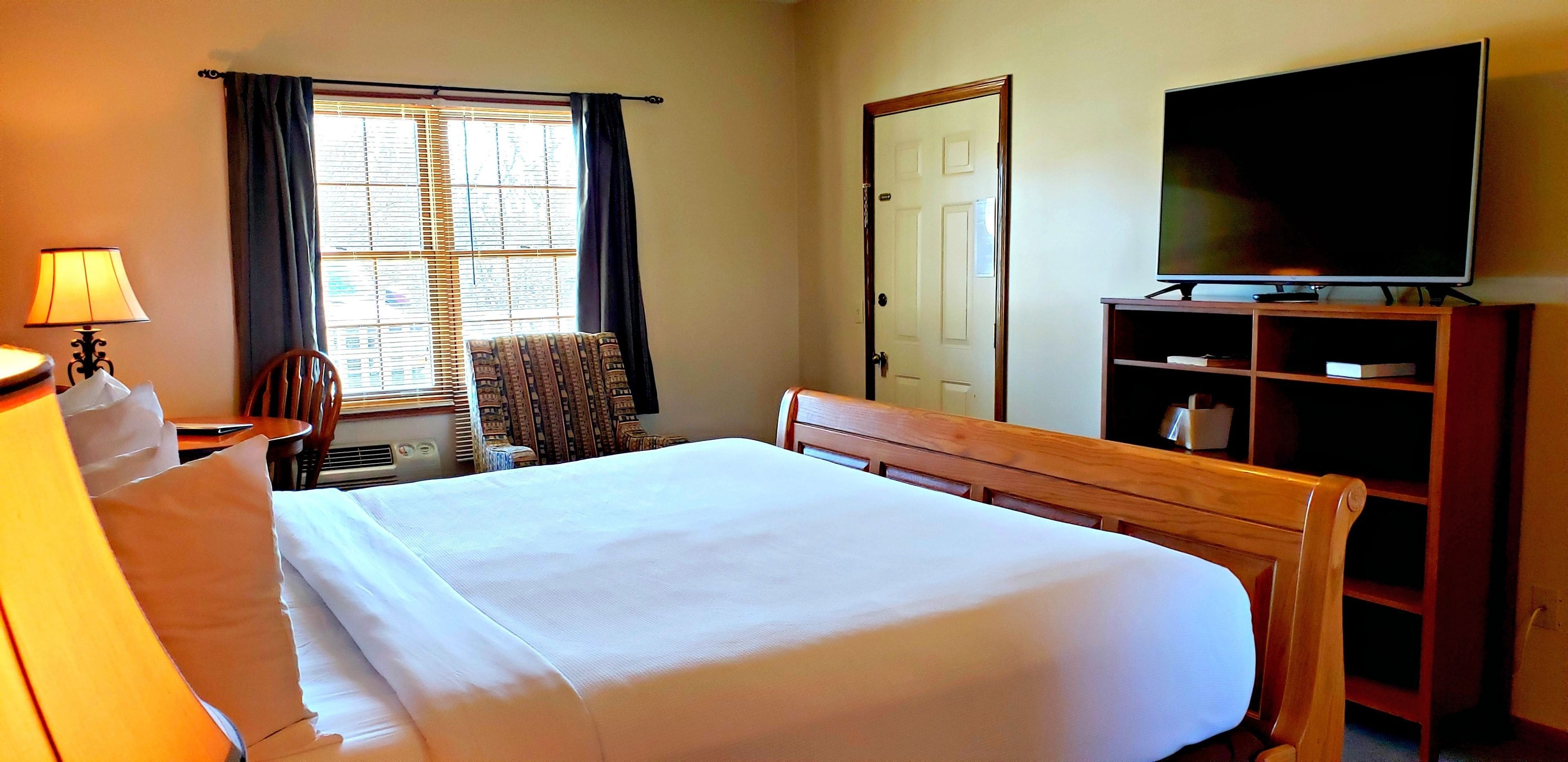 Een hotelkamer met een bed, een bureau met lamp, een stoel, een televisie op een standaard en een raam met gordijnen.