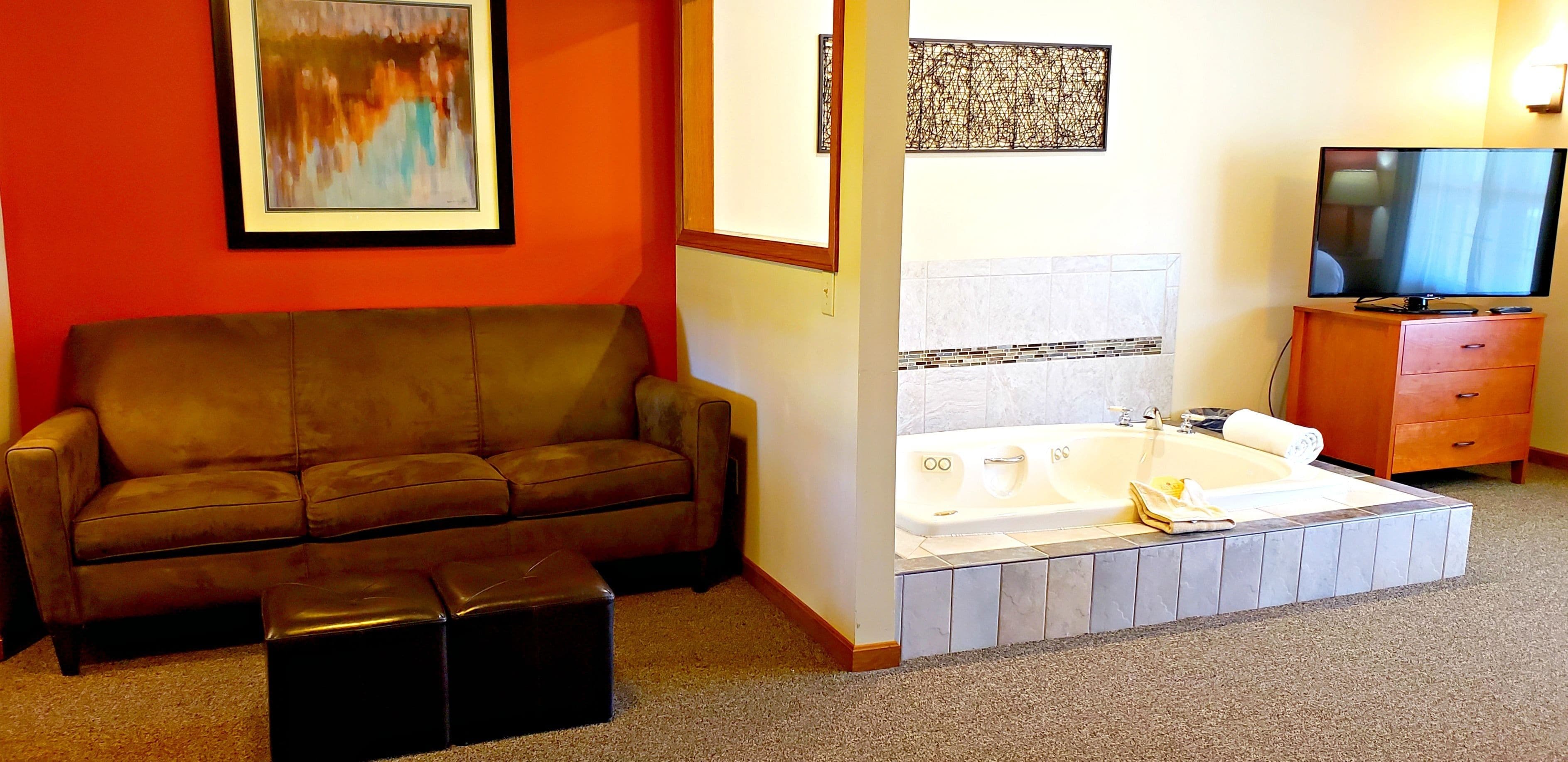 A room with a brown leather sofa, a black ottoman, a white bathtub, a wooden cabinet, and a television.