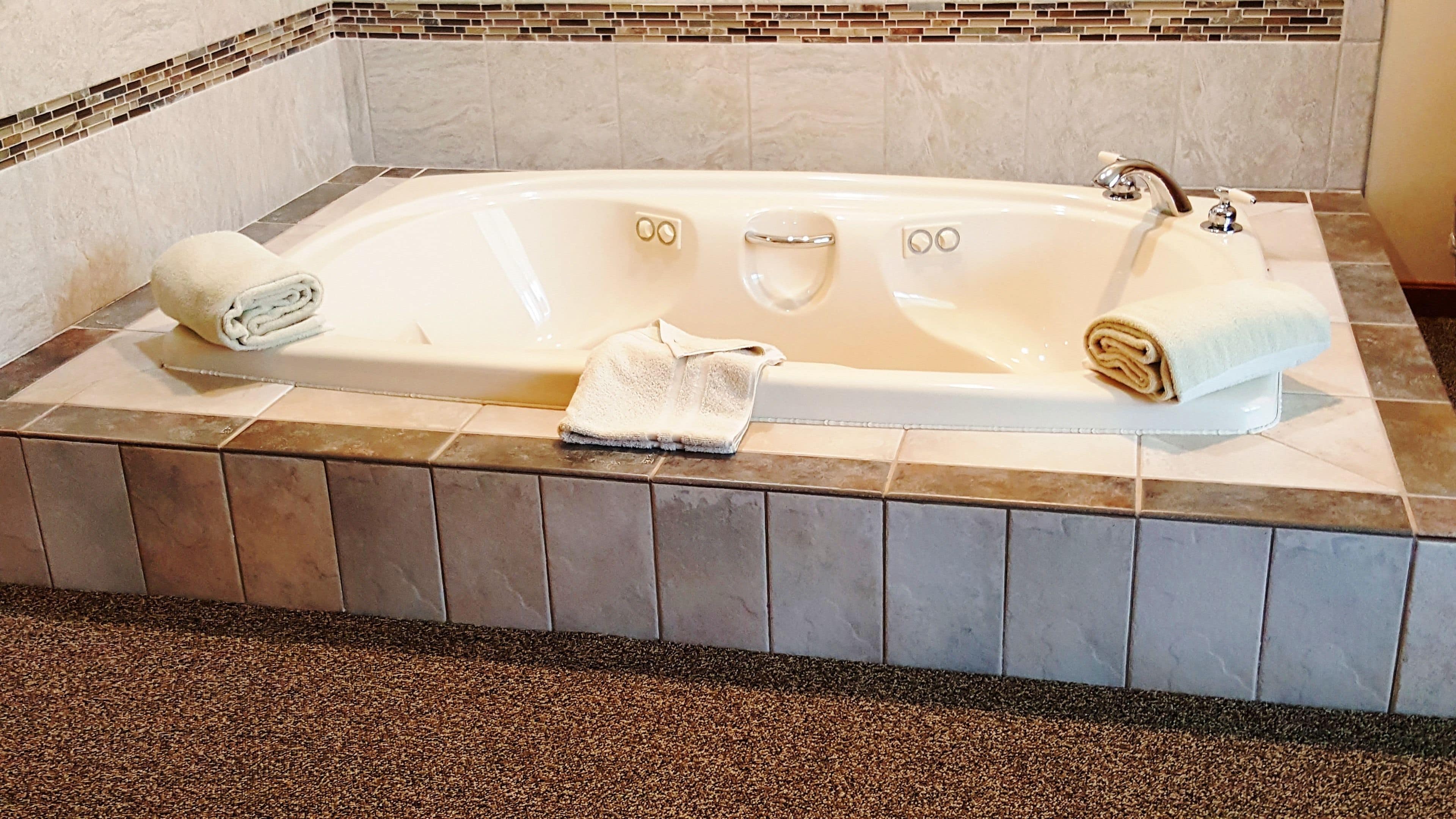 A large, rectangular bathtub with two faucets, a built-in seat, and a small control panel. There are two towels, one on each side of the tub. The floor is tiled with a mix of brown and beige tiles, and the walls have a mosaic tile border.
