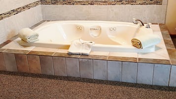 A large, rectangular bathtub with two faucets, a built-in seat, and a small control panel. There are two towels, one on each side of the tub. The floor is tiled with a mix of brown and beige tiles, and the walls have a mosaic tile border.