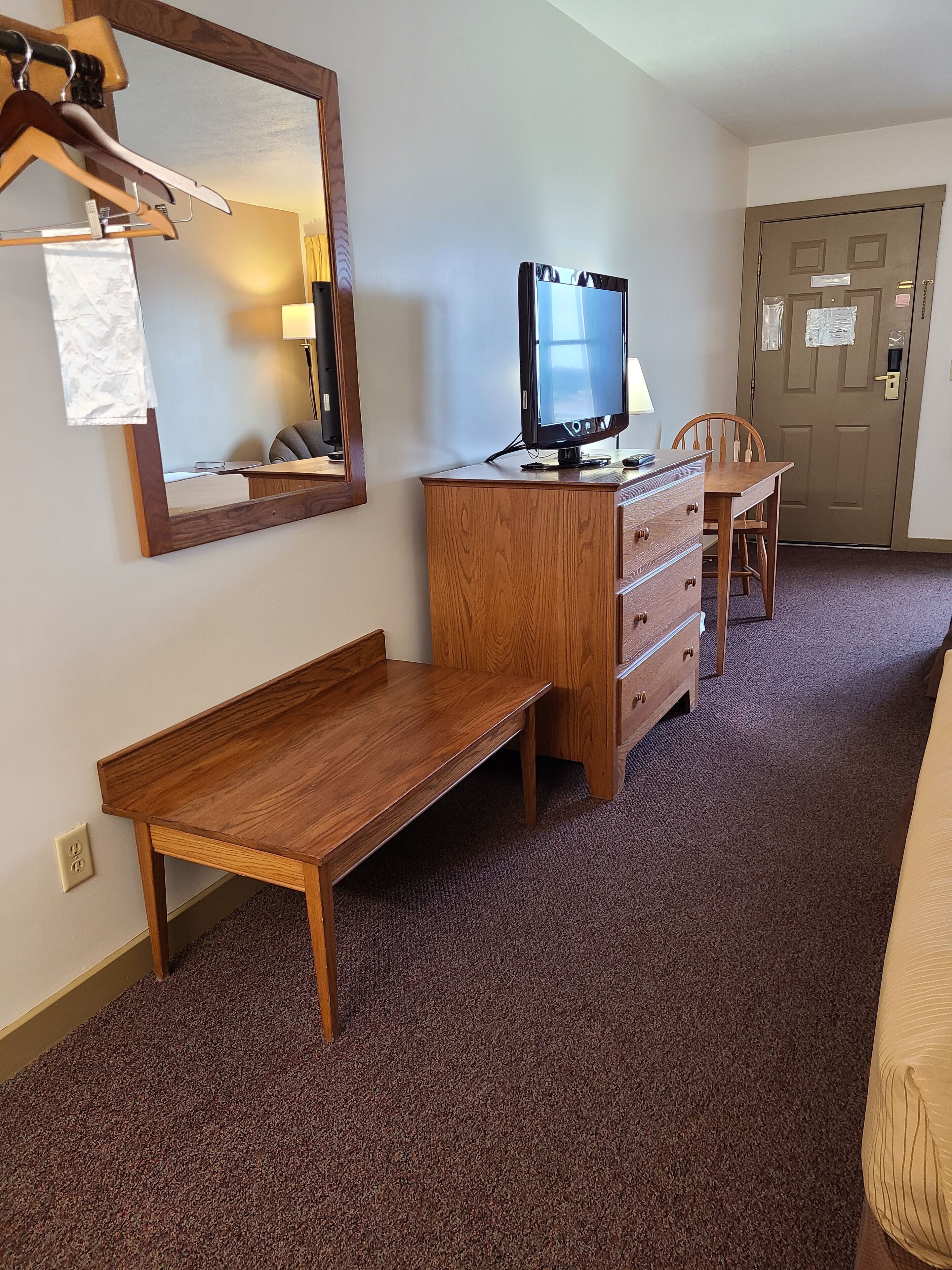 A hotel room with a wooden desk, a dresser, a chair, a television, and a door.