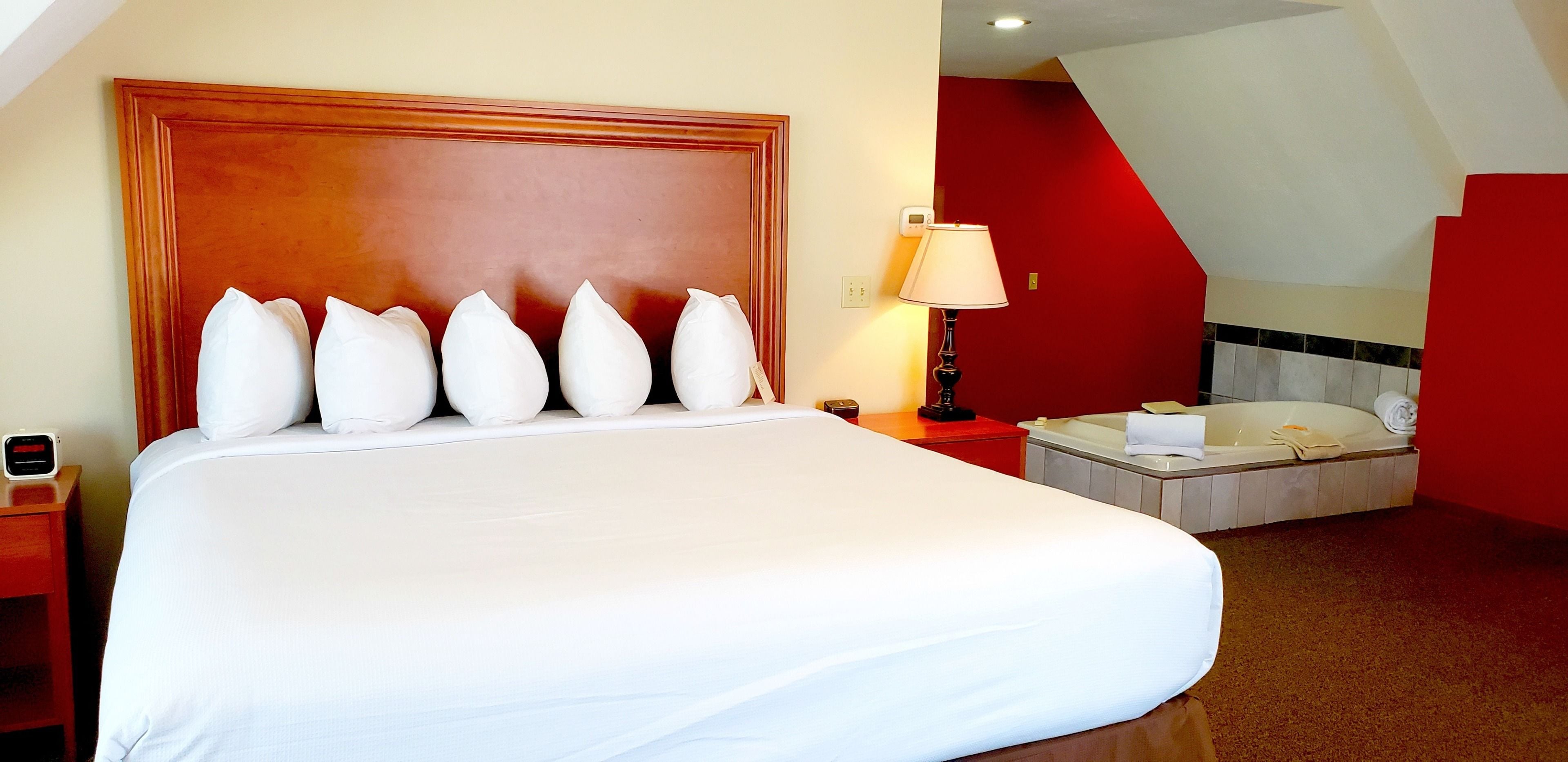 A hotel room with a large bed, a bedside table with a lamp, a red wall, and a bathtub.