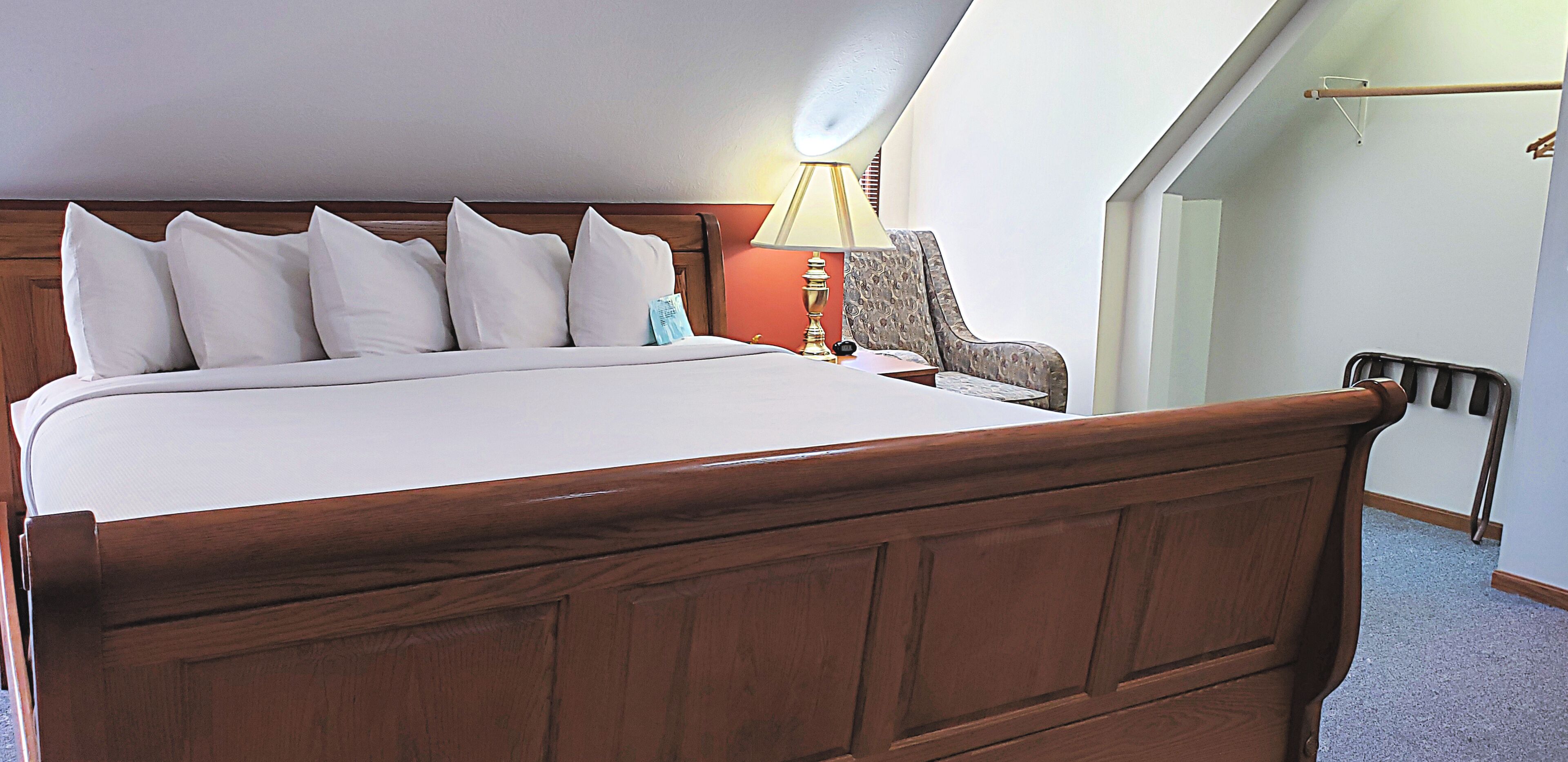 A neatly made bed with a wooden headboard, a bedside lamp, and a chair in a room with a sloped ceiling and a visible door.