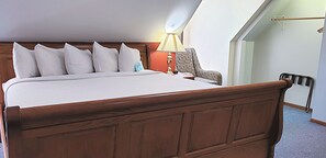 A neatly made bed with a wooden headboard, a bedside lamp, and a chair in a room with a sloped ceiling and a visible door.
