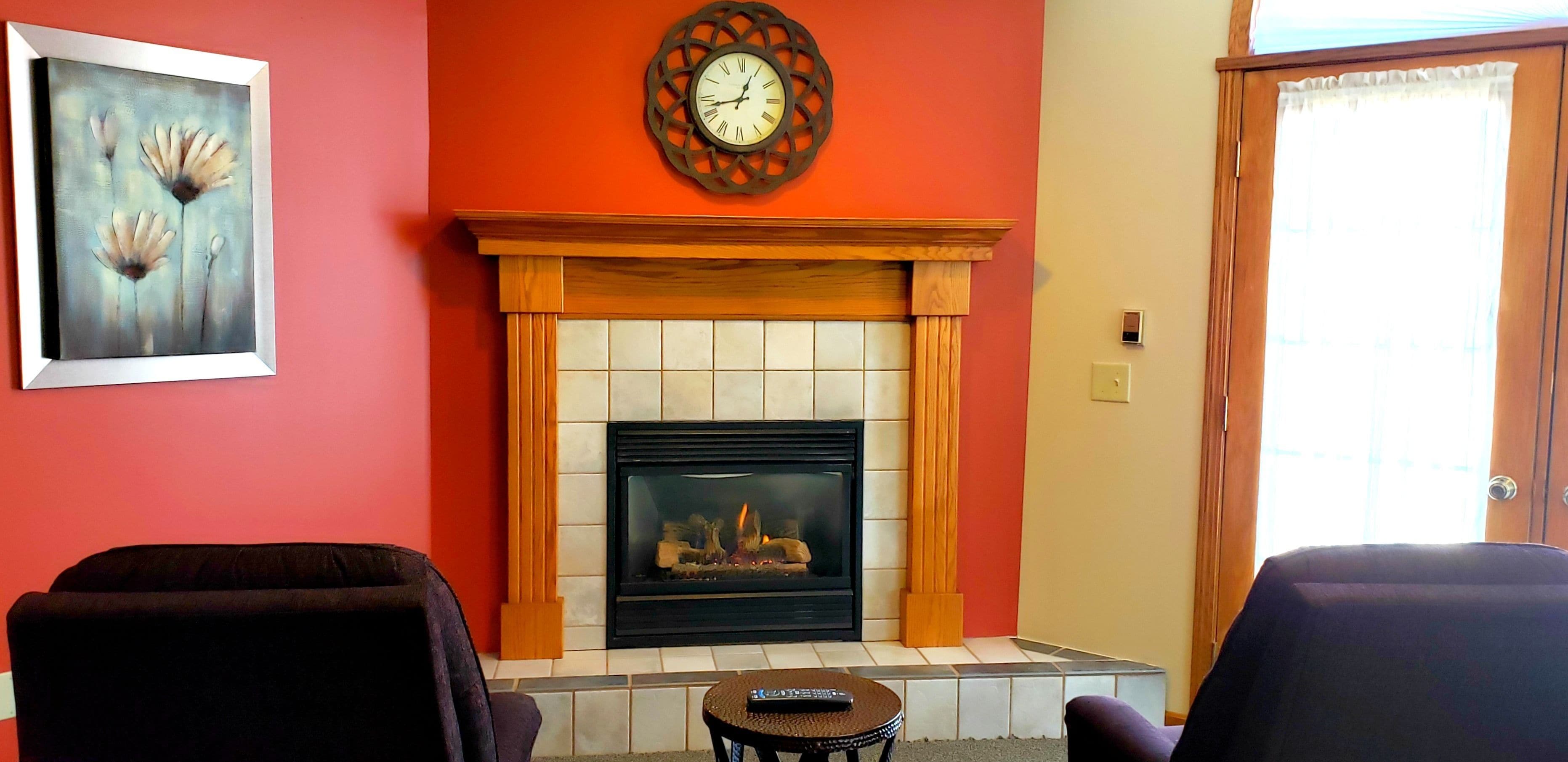 A living room with a fireplace, two armchairs, a small round table, and a framed painting on the wall.