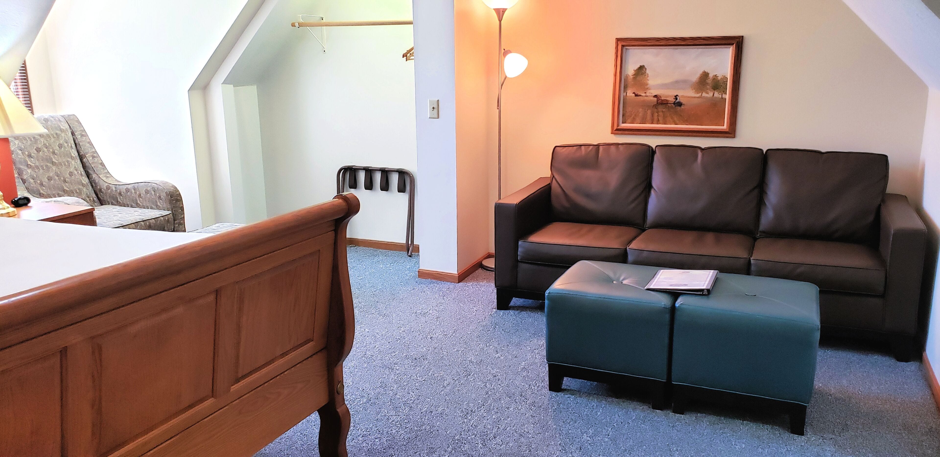 A room with a wooden bed, a brown leather sofa, a green ottoman, a framed picture on the wall, and a floor lamp.